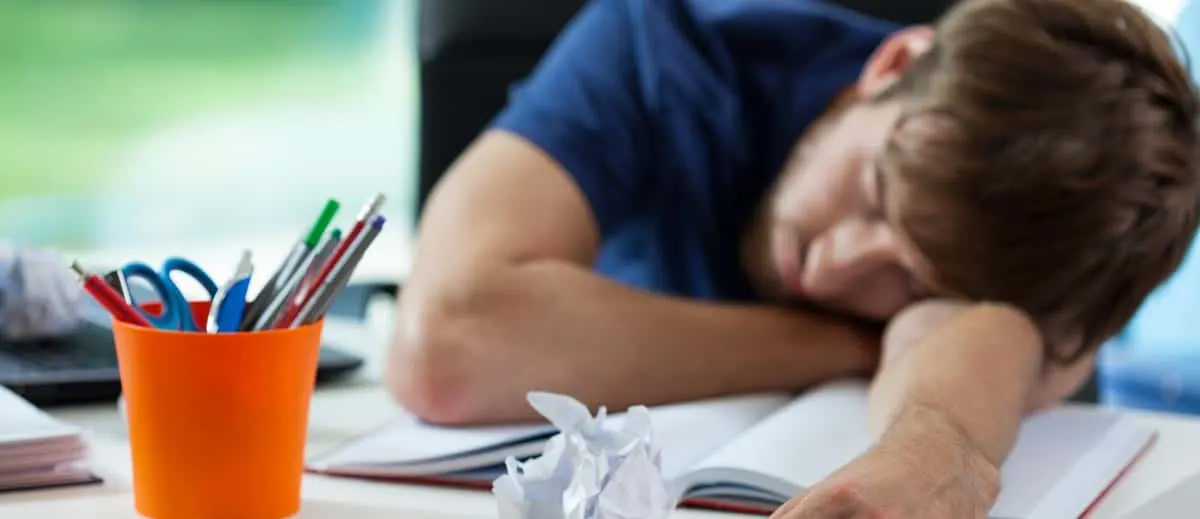 Young man asleep at his desk with books and papers scattered, representing fatigue and poor focus from drug use consequences.
